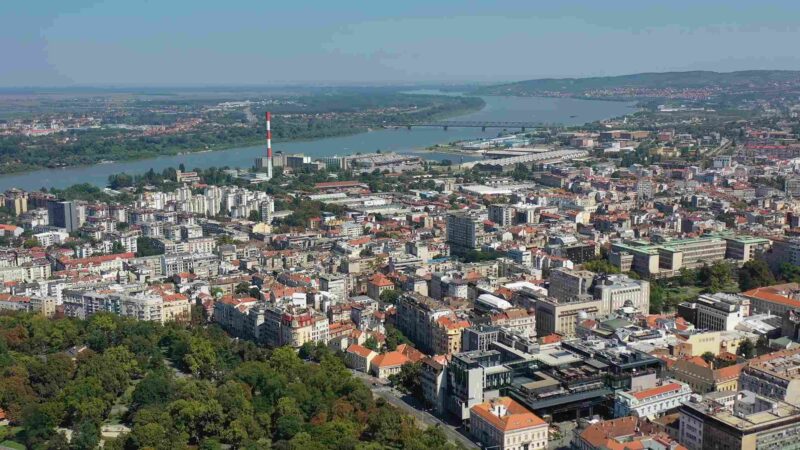 Aerial view of Belgrade with city buildings, bridges, green areas, and rivers in the background.
