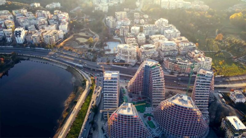 Aerial view of modern city buildings, roads, parkland, and a lake at sunset.