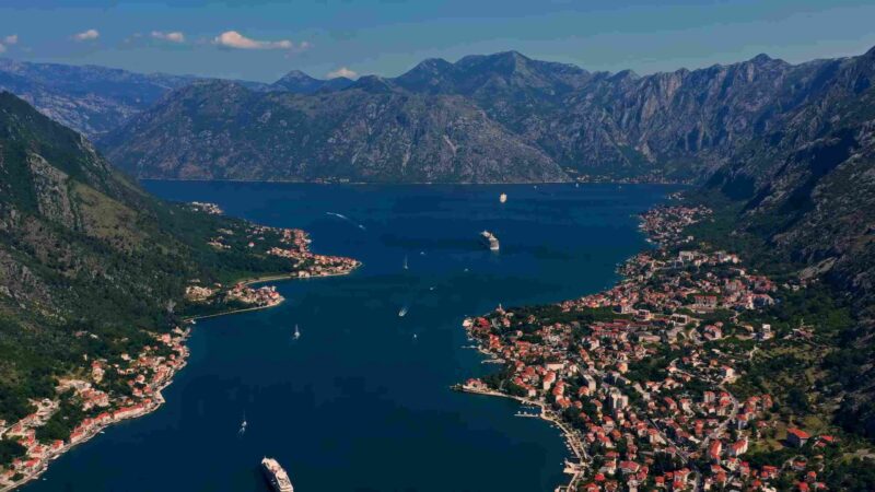 Aerial view of Kotor Bay with blue water, coastal towns, boats, and rugged mountains.