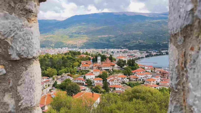 View of Ohrid with red-roofed houses, churches, mountains, and Lake Ohrid seen through stone walls.