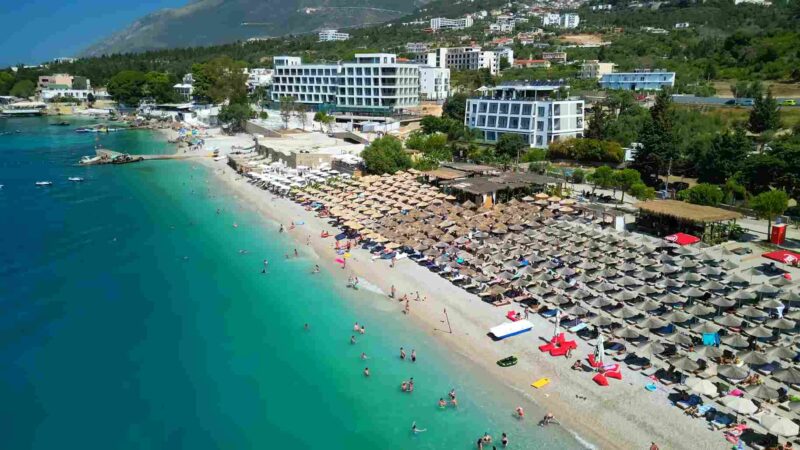 Aerial view of a busy Albanian Riviera beach with clear turquoise water, sun umbrellas, hotels, and mountains.