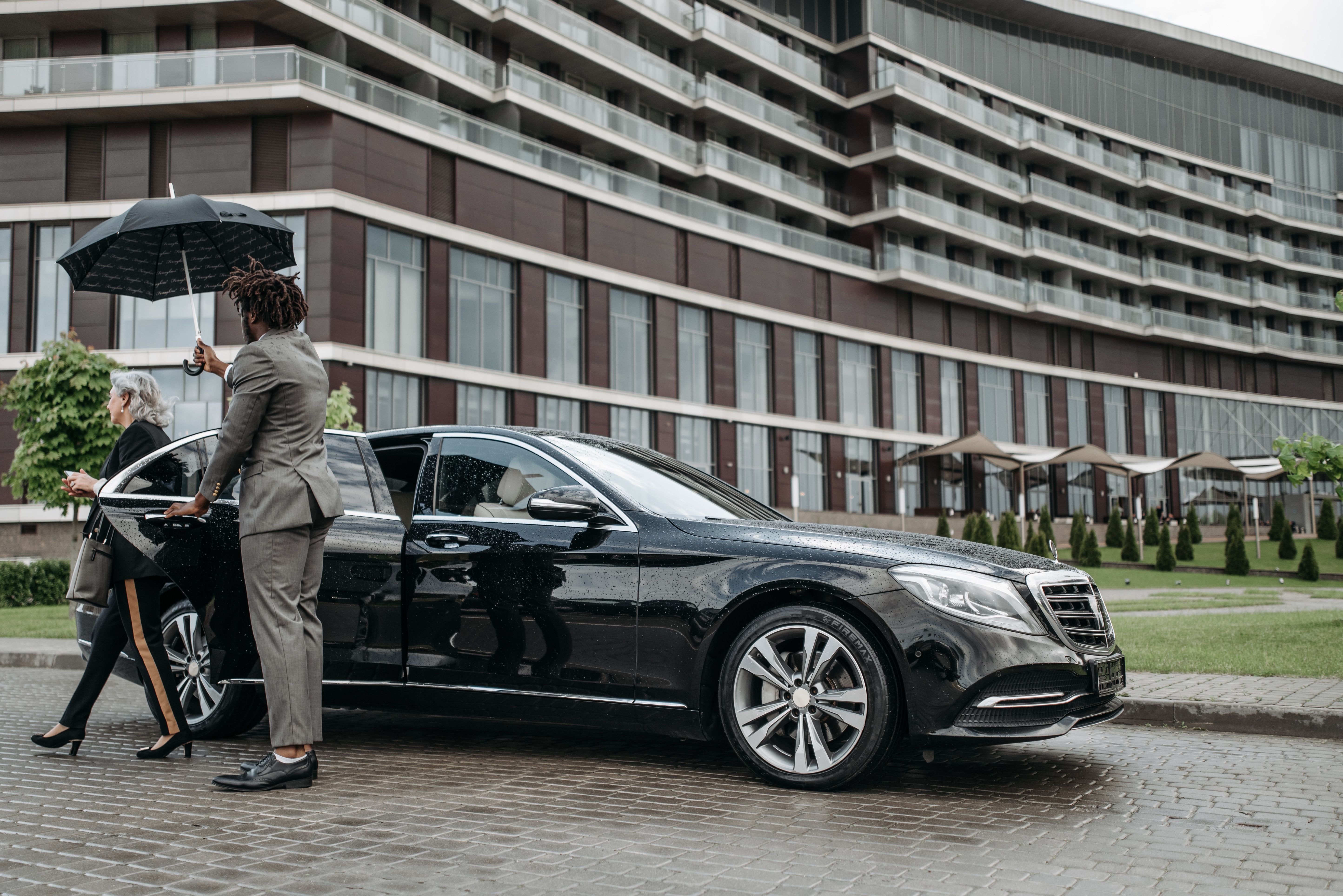 A traveler is shielded by an umbrella while exiting a luxury car, suggesting privacy and discretion.