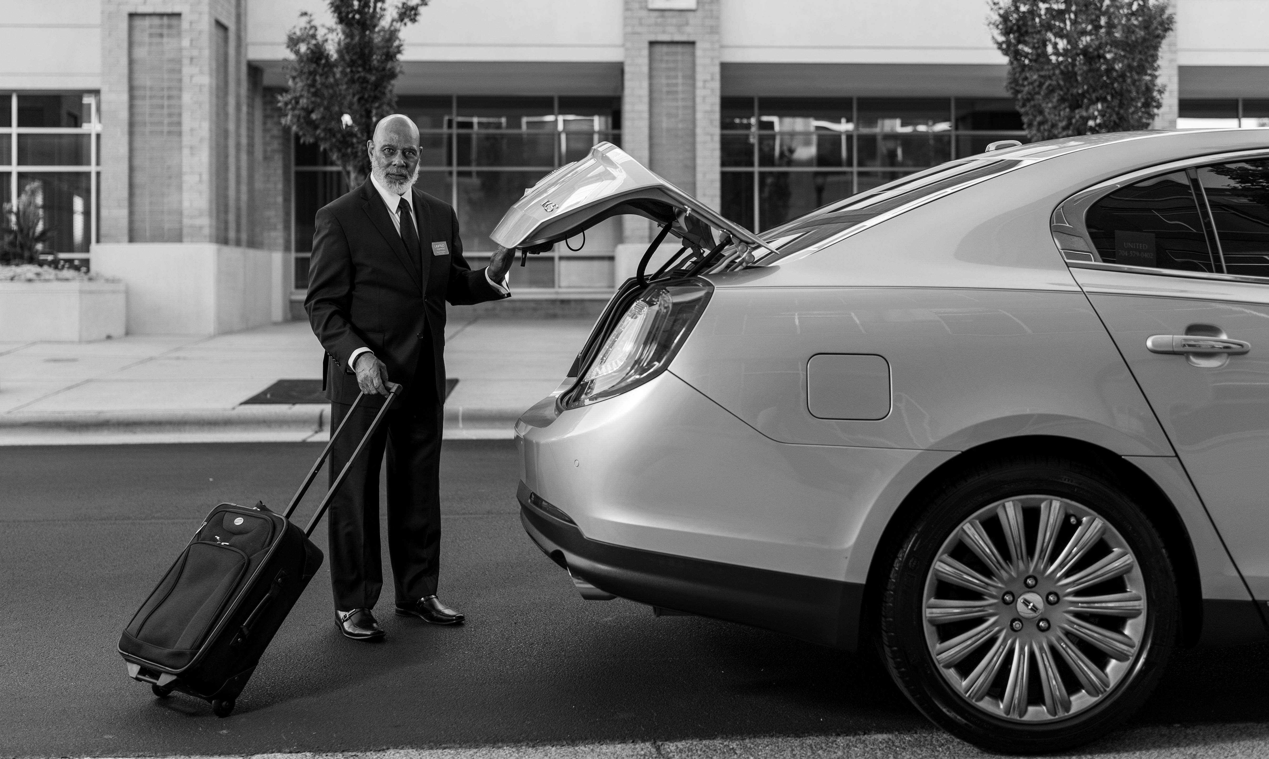 Chauffeur discreetly loading luggage into a luxury car outside a modern building.